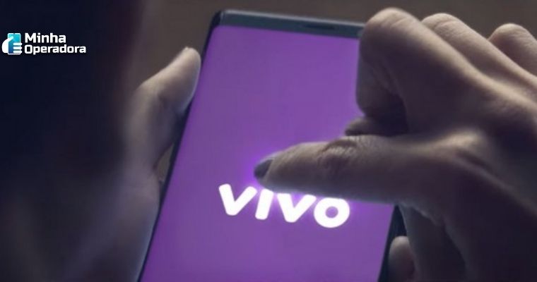 Pessoa utilizando o celular, enquanto na tela aparece a logomarca da Vivo na cor branca com o fundo roxo.