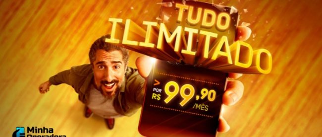 Marcos Mion levanta um celular que traz a oferta na tela.