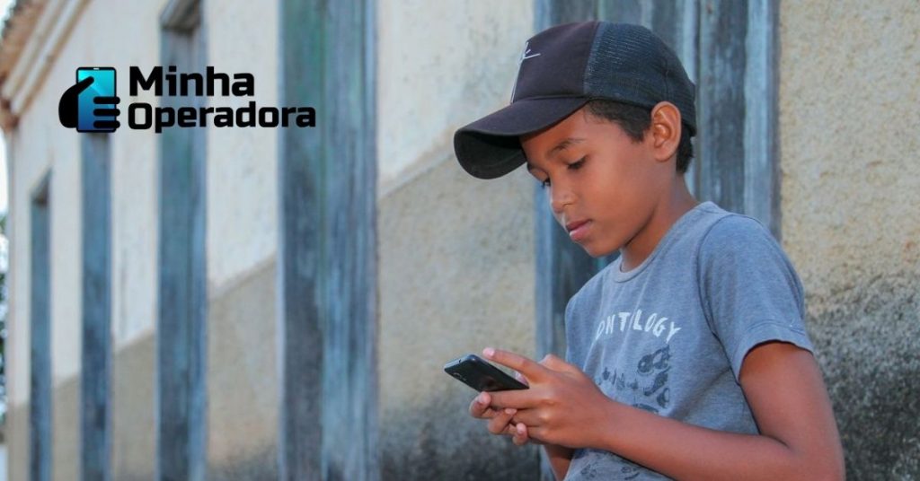 Menino de boné utilizando o celular na frente de casa.