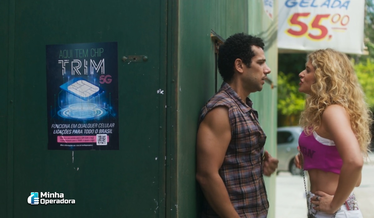 A imagem mostra dois personagens da novela Volta por Cima em frente a uma parede verde. Na parede, há um cartaz anunciando o "TRIM 5G", que é um chip de celular. O cartaz diz: "Aqui tem chip TRIM 5G" e menciona que o chip funciona em qualquer celular e permite ligações para todo o Brasil. Também há um QR code no cartaz para mais informações. À direita dos personagens, há uma placa ao fundo que anuncia "Cerveja Gelada" por R$ 55. A cena mostra uma conversa casual entre os personagens.