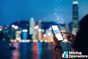 Pessoa utilizando telefone que tem a inscrição "5G" na tela. Ao fundo, o horizonte de uma cidade à noite.