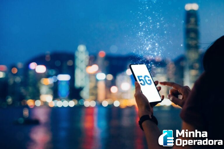 Pessoa utilizando telefone que tem a inscrição "5G" na tela. Ao fundo, o horizonte de uma cidade à noite.